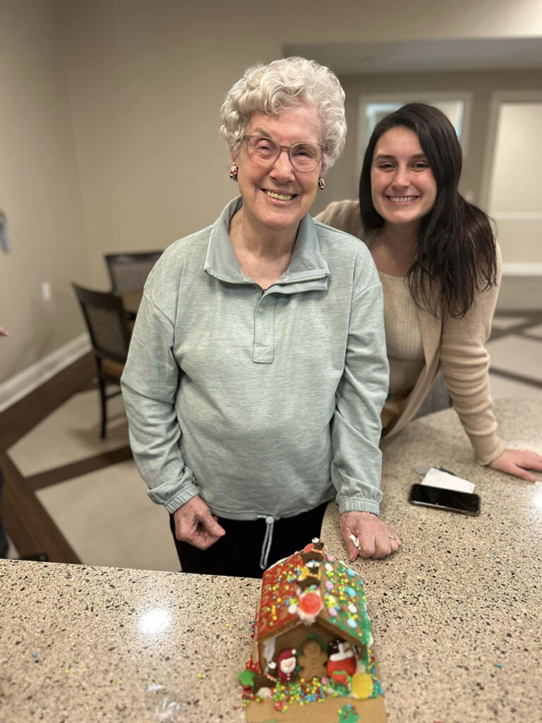 Witness the joy of collaboration as an elderly woman and a young lady stand beside their beautifully decorated gingerbread house.