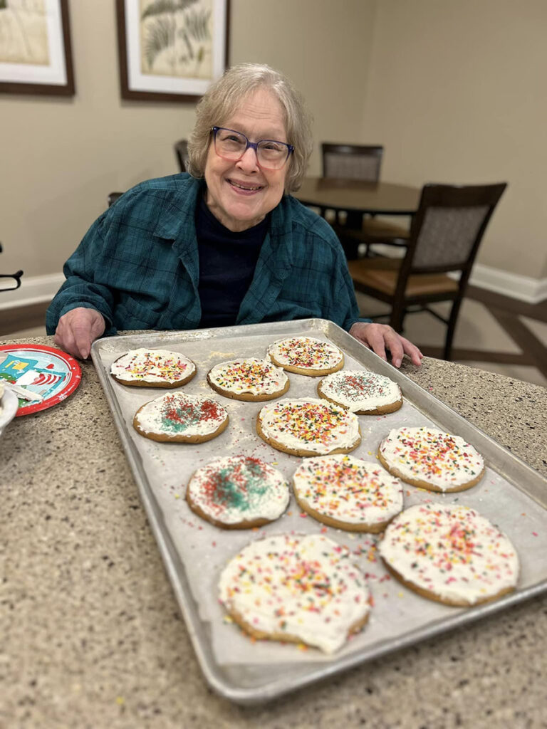 A delightful community resident, wearing a teal plaid long sleeve, beams with joy while presenting a tray of deliciously frosted and sprinkled cookies.