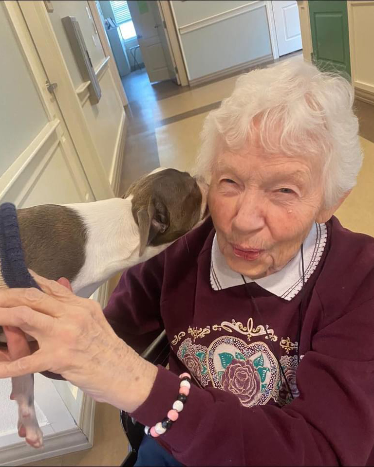 Elderly woman in wheelchair smiling, holding a dog brought by a volunteer for memory care residents.