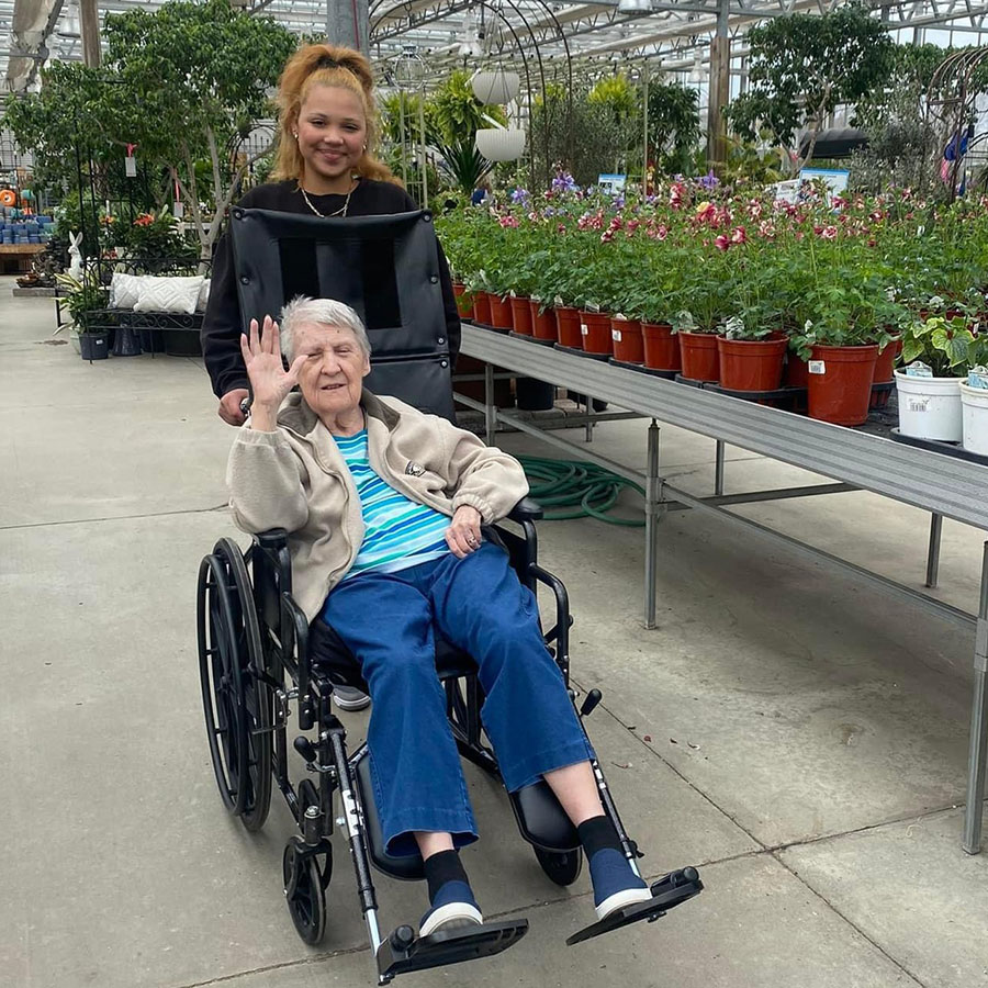 Volunteer assisting elderly woman in wheelchair at greenhouse, gathering flowers.