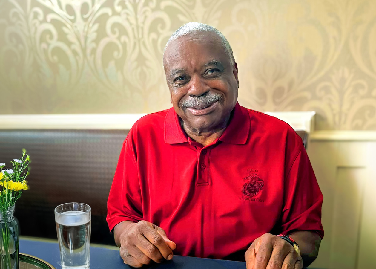 Smiling senior man wearing a red polo shirt, sitting at a table with a glass of water and a vase of flowers, indoors.