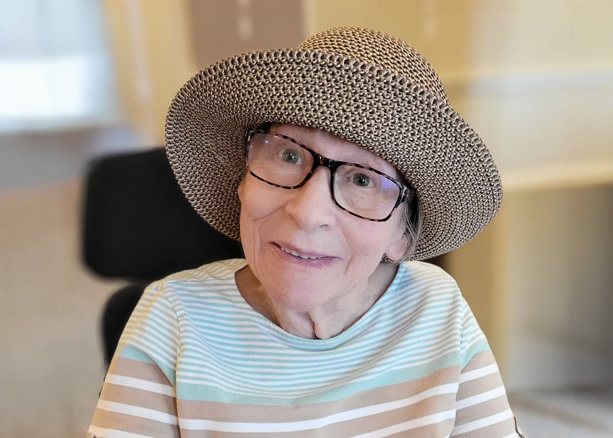 A smiling senior lady wearing glasses and a wide-brimmed hat sits indoors with a striped shirt, attending an ice cream Happy Hour.