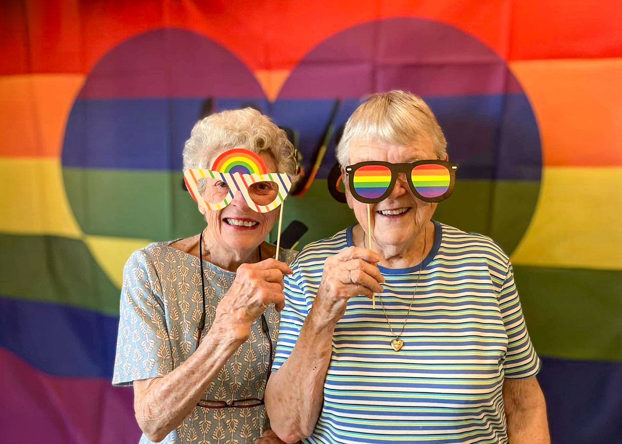 Two senior ladies smiling and holding rainbow-themed props in front of a colorful background during Pride Week celebrations.