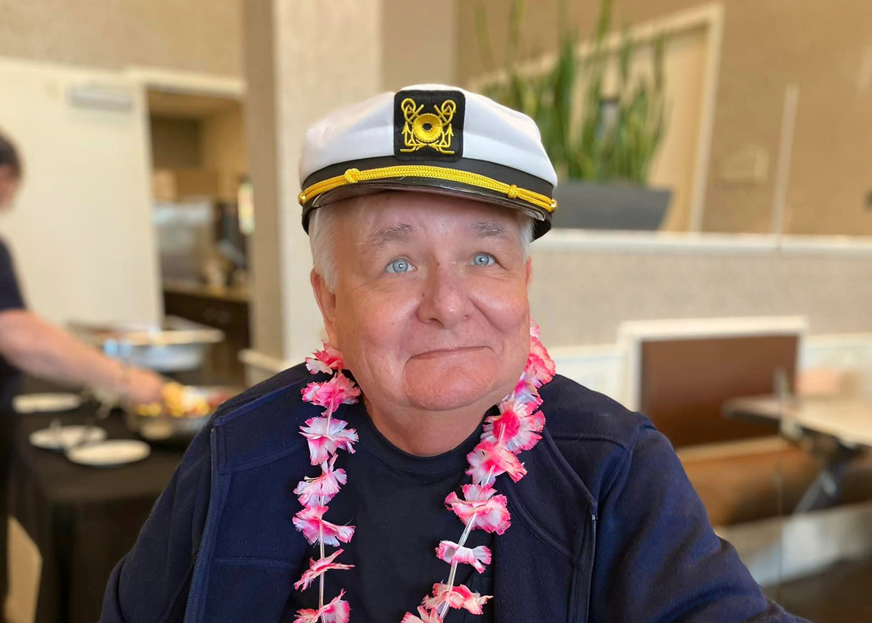 Smiling senior man wearing a captain's hat and a pink lei, sitting indoors during a themed event.