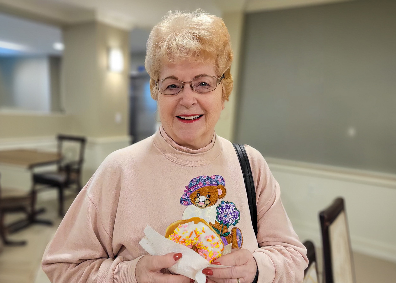 Smiling senior lady wearing glasses and a sweater with a bear design, holding a donut with colorful sprinkles indoors.