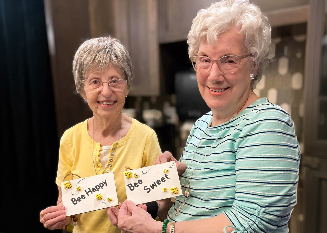 Two senior ladies smiling and holding up handmade signs that read 'Bee Happy' and 'Bee Sweet' indoors.