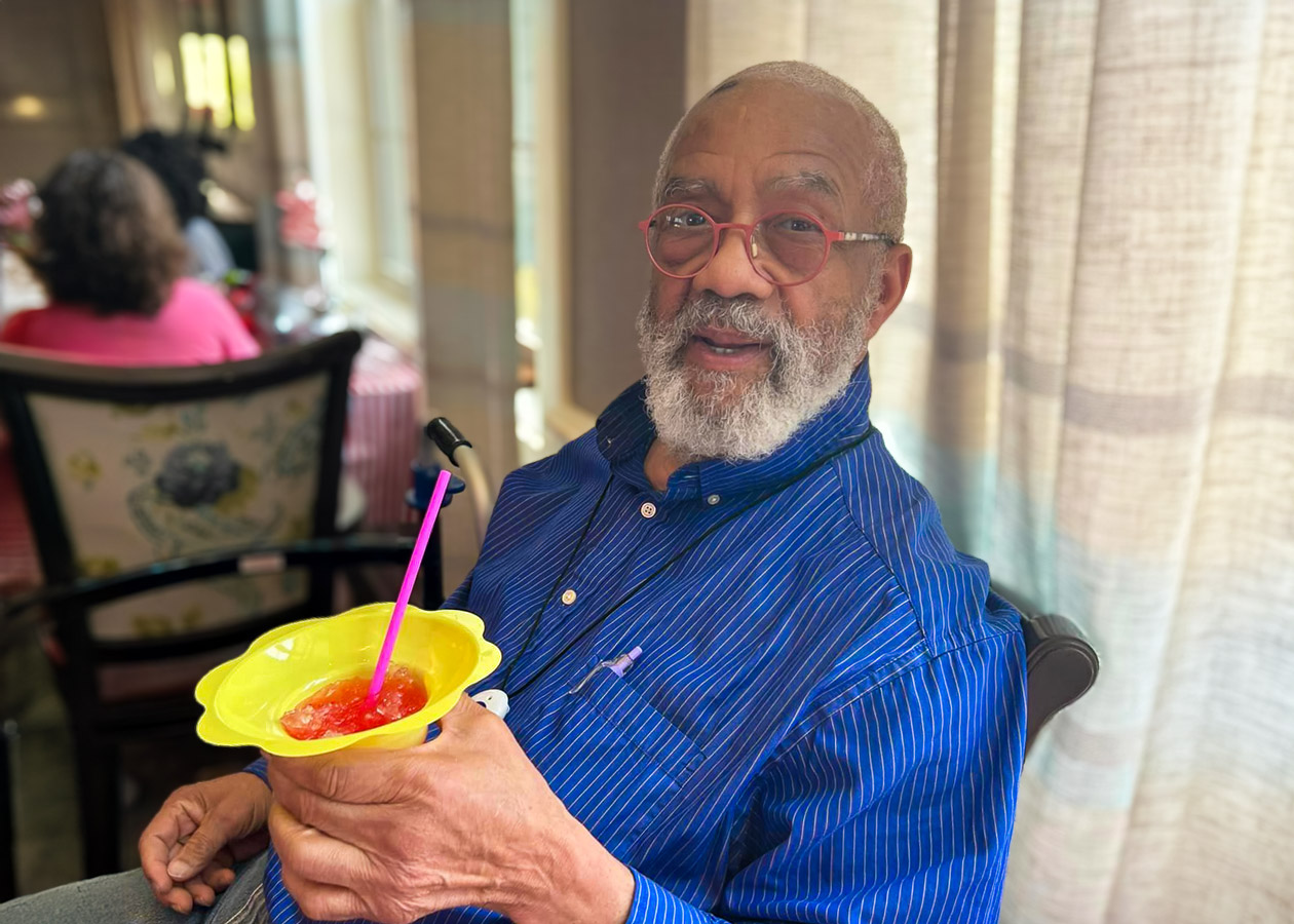 Smiling senior man with a beard and glasses, wearing a blue shirt, holding a yellow cup with a pink straw, sitting indoors.