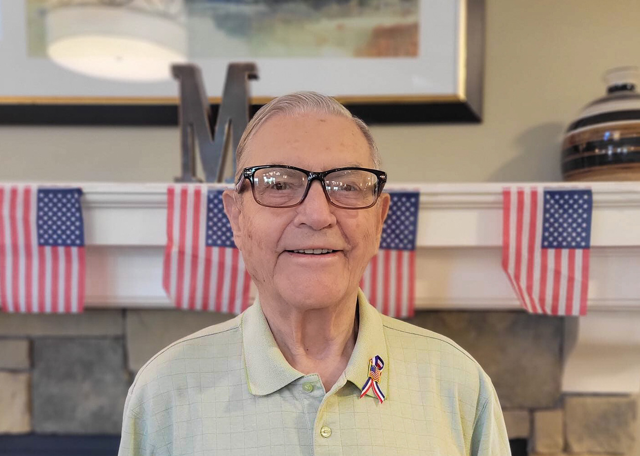 Smiling senior man wearing glasses and a light green polo shirt with an American flag pin, standing indoors with a patriotic backdrop.