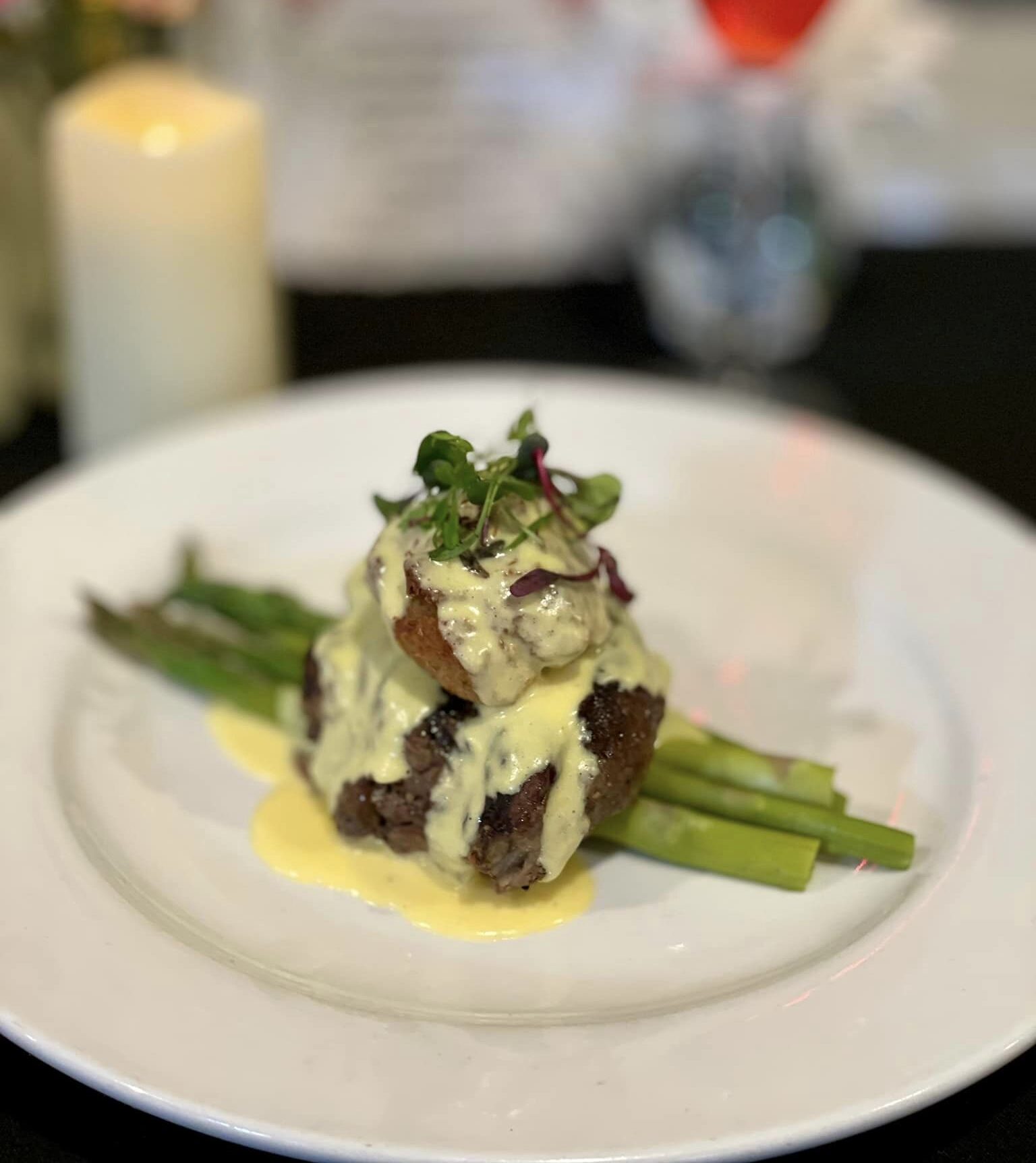 Plated gourmet steak with asparagus, topped with creamy hollandaise sauce and garnished with microgreens, set against a blurred background of a candle, flowers, and drink.
