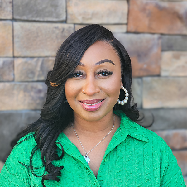 Melody Gordon, Operations Coordinator at The Madison Senior Living, smiles warmly in a vibrant green blouse against a stone wall, reflecting her dedication to senior living excellence.