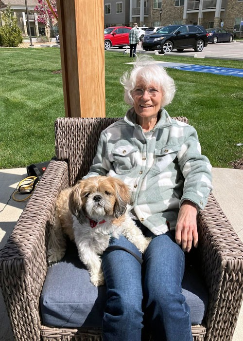 A senior resident relaxes in a wicker chair with a small dog on her lap, enjoying the sunshine outside at The Madison Senior Living.