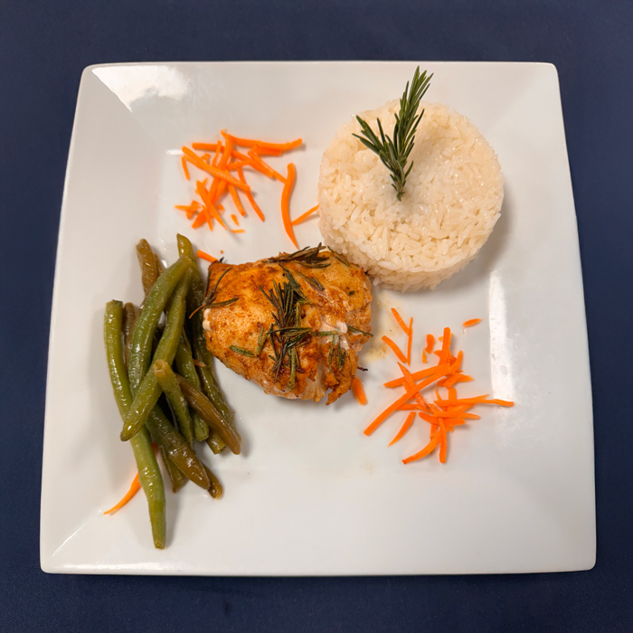 Plated meal featuring rosemary-seasoned chicken, sautéed green beans, shredded carrots, and a molded serving of white rice topped with a rosemary sprig.
