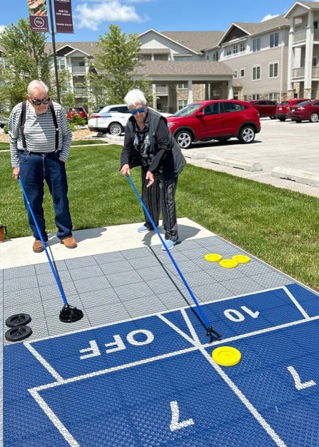 Two senior residents play a game of outdoor shuffleboard on a bright spring day at The Madison Senior Living, with the community building in the background.