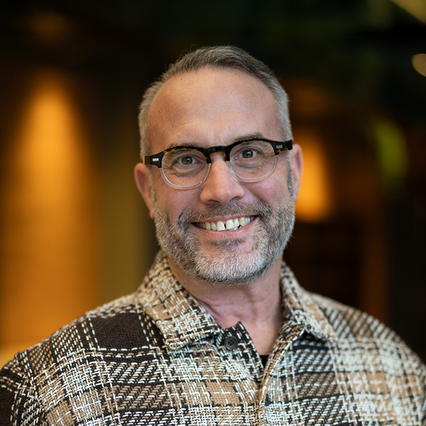 Todd Bettey-Donelson, Executive Director at The Madison Senior Living, smiling in a warmly lit indoor setting and wearing glasses and a plaid-patterned shirt.