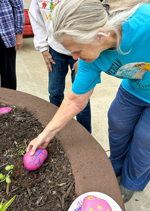 Senior resident places a brightly painted rock in a garden bed during a community art project, adding color and creativity to the outdoor courtyard space.