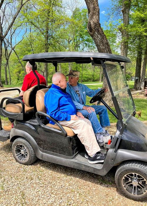 Senior residents enjoy a peaceful outing, riding together in a golf cart through a scenic wooded area on a sunny day filled with nature and conversation.
