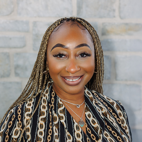 Melody Gordon, Operations Coordinator at The Madison Senior Living, smiling in front of a gray stone wall, wearing a patterned blouse and layered necklaces.