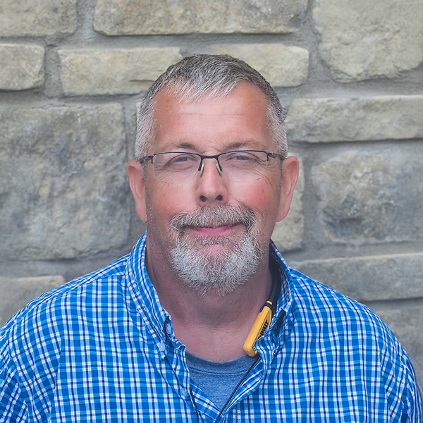Keith Miller, Plant Operations Director at The Madison Senior Living, smiles in front of a stone wall while wearing a blue plaid shirt and glasses.