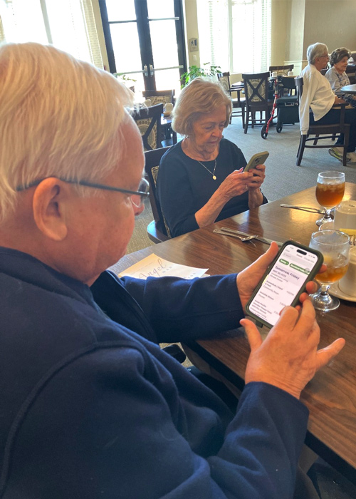 A senior man and woman seated at a dining table explore the Arrow app on their smartphones, reviewing the menu and community options during a meal.