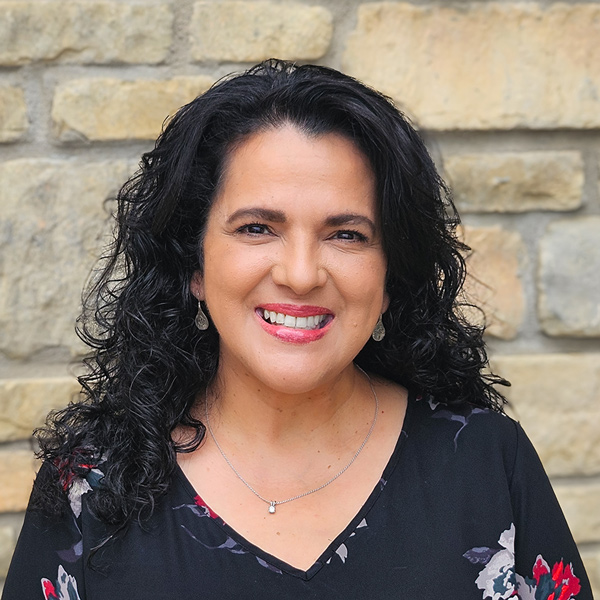 Nancy Majors, Director of Wellness at The Madison Senior Living, standing in front of a stone wall, smiling, with curly black hair and wearing a black floral top.