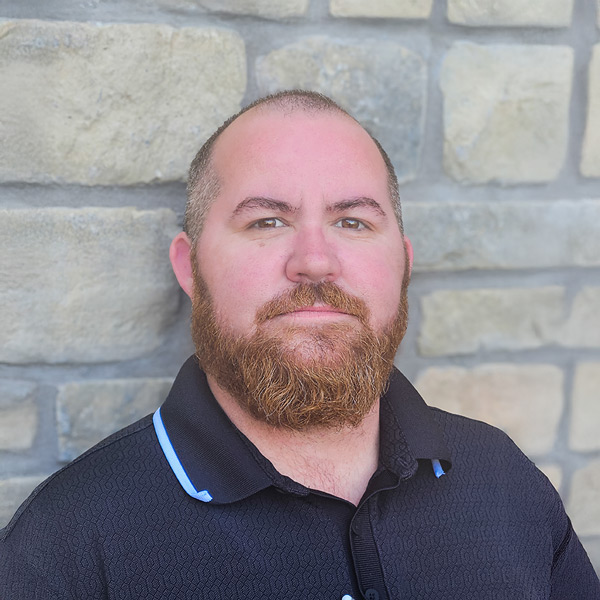 Tyler Sproston, Culinary Director at The Madison Senior Living, standing in front of a stone wall and wearing a black collared shirt with light blue trim.