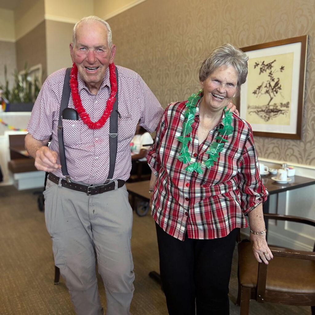Smiling senior couple wearing leis enjoy a festive celebration at the community, sharing laughter and joy during a themed social gathering.