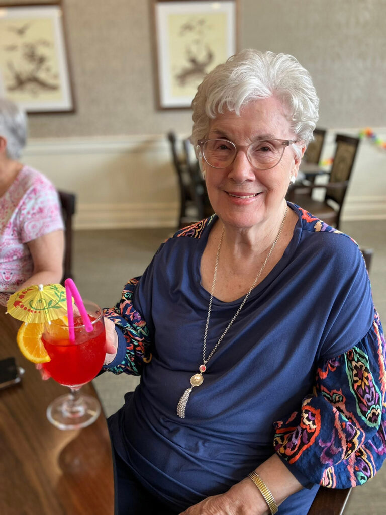 Senior woman with glasses smiles while holding a bright red tropical drink with fruit and umbrella garnish at a community luau-style event.
