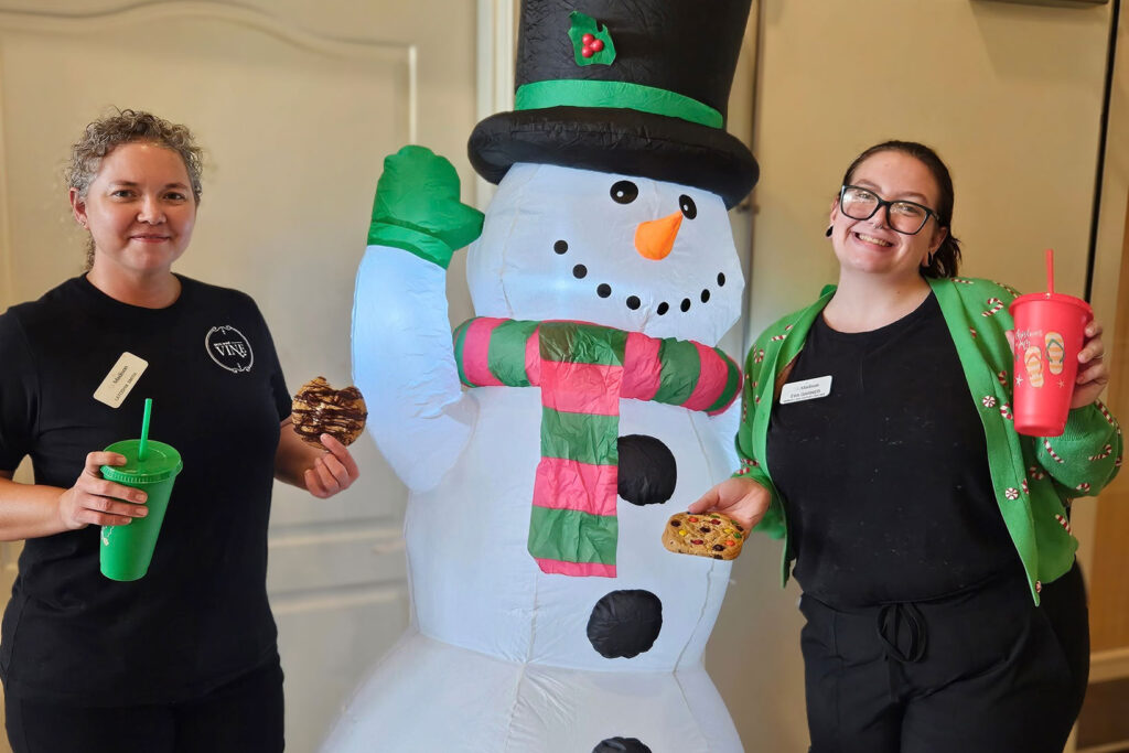 Team members smile beside a festive inflatable snowman, holding colorful holiday drinks and cookies, celebrating the season with cheer at the community.