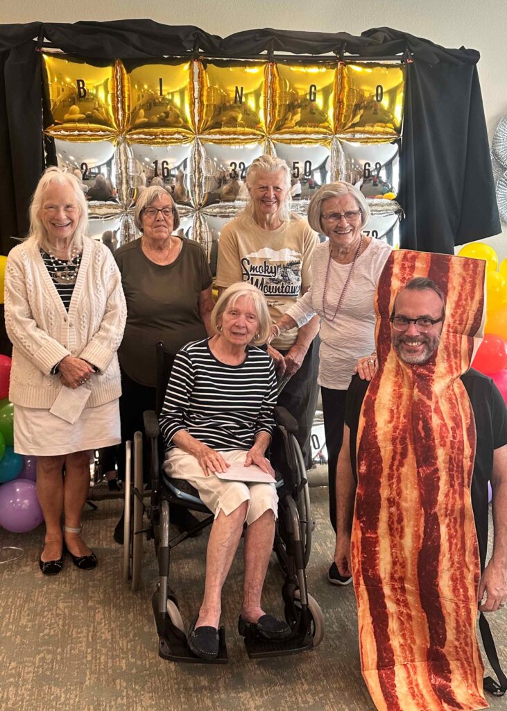 A group of residents pose in front of a shiny gold and silver bingo backdrop, smiling with a staff member dressed in a playful bacon costume during a themed event.