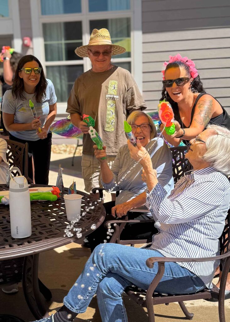 Residents and team members laugh together outside, playfully holding colorful water blasters during a fun summer celebration in the senior living courtyard.