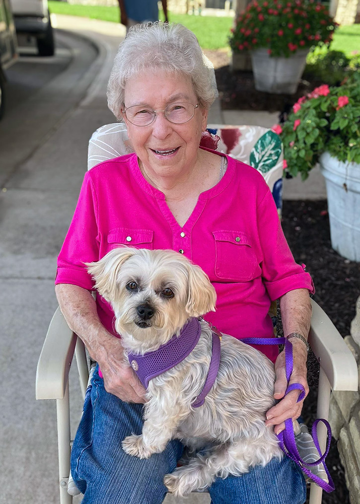 A senior woman in a bright pink shirt sits outside in a patio chair with a fluffy white dog on her lap.