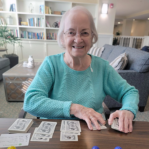 A senior woman smiles brightly, shuffling cards at The Madison Senior Living.