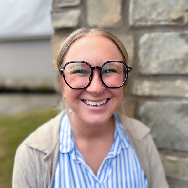 Brittny Middleton, Memory Care Director at The Madison Senior Living, smiling in a professional headshot, wearing glasses and a striped blouse, with a softly blurred stone and outdoor background.