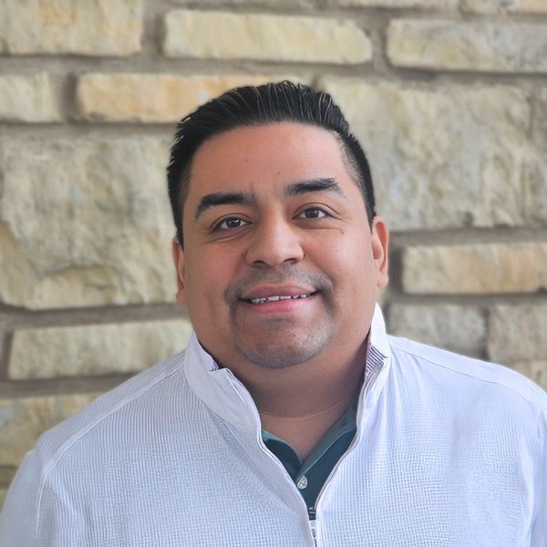 Jesse Guillen, Senior Living Director at The Madison Senior Living, smiling in a professional headshot, wearing a light quarter-zip jacket, with a stone wall background.