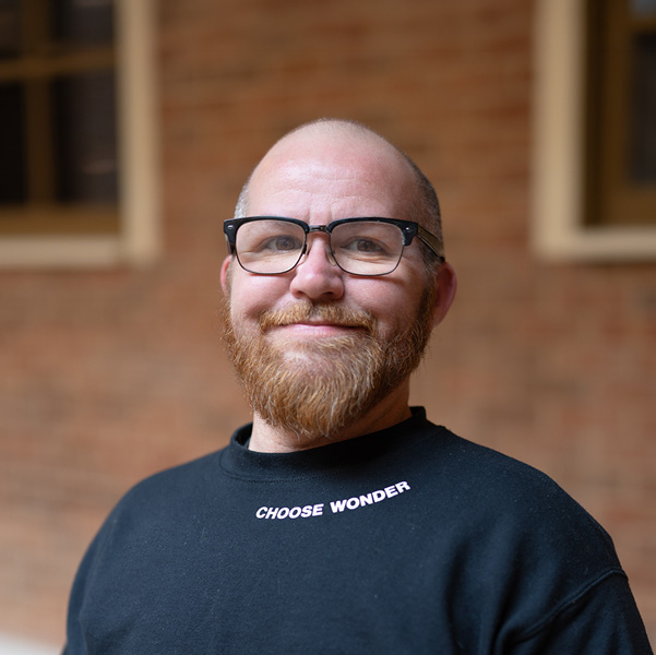 Tyler Sproston, Culinary Director at The Madison Senior Living, smiling in a professional headshot, wearing glasses and a black shirt, with a softly blurred brick and window background.