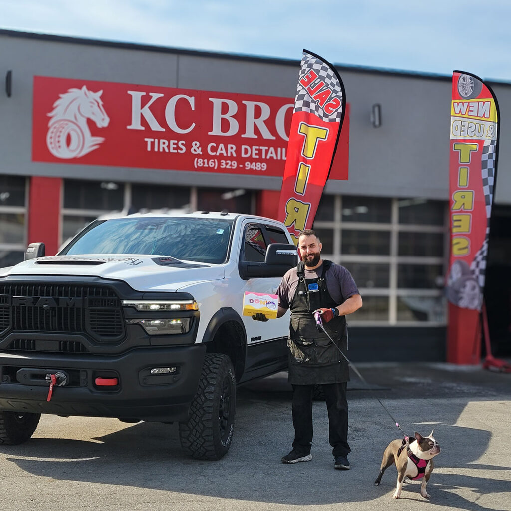 A man at KC Bros Tires & Car Detail smiles in front of his business and truck, holding a box of gifted donuts in one hand and the leash to his canine companion in the other.