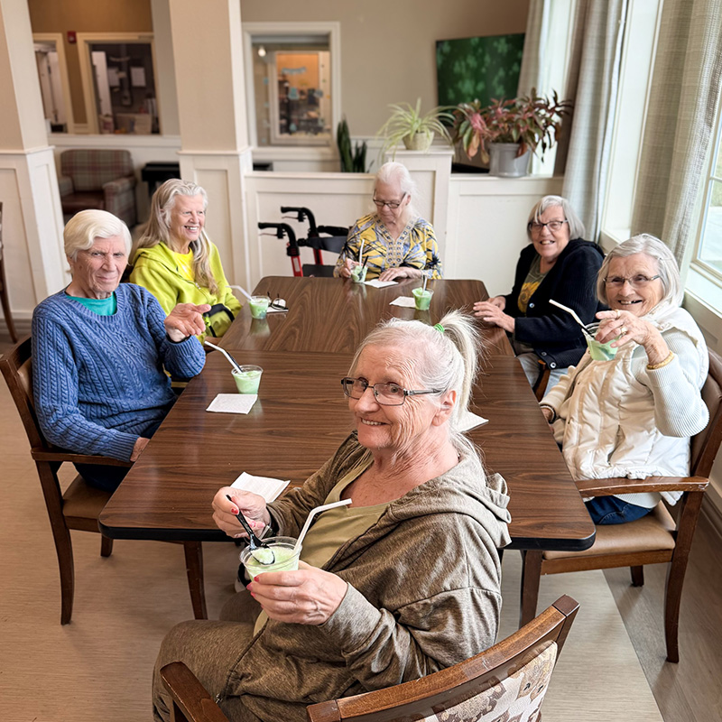 A group of residents gather around a table at The Madison Senior Living, smiling in relaxed conversation.