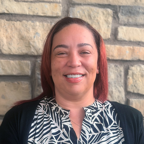 Latrisha West, Wellness Director at The Madison Senior Living, smiling in a professional headshot with straight red-toned shoulder-length hair, a patterned black and white blouse, and a light stone wall background.