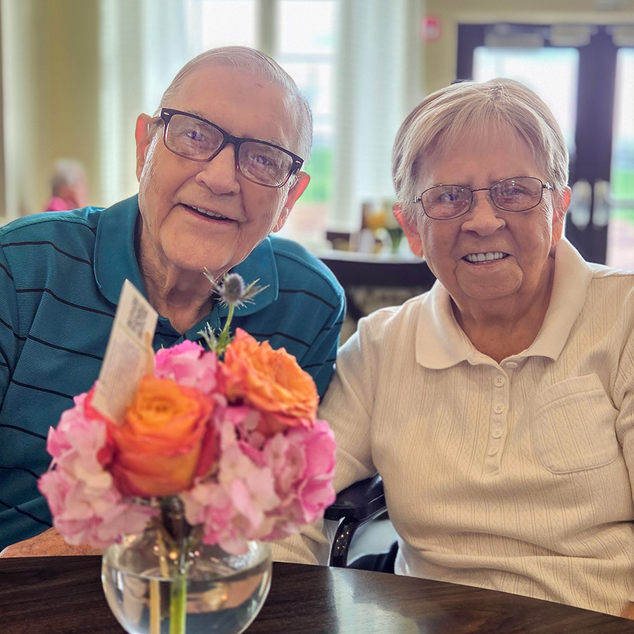 Two senior residents at The Madison Senior Living smile brightly beside a bouquet of flowers.