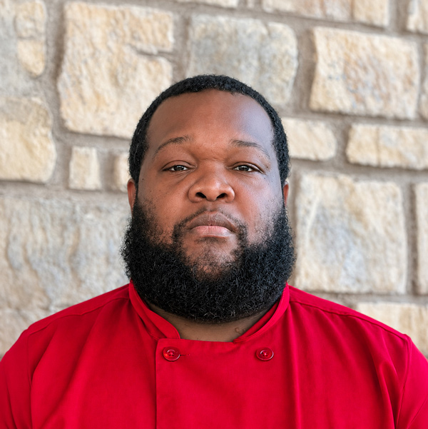 Albert Wright, Culinary Director at The Madison Senior Living, in a professional headshot wearing a bright red chef coat before a light stone wall.