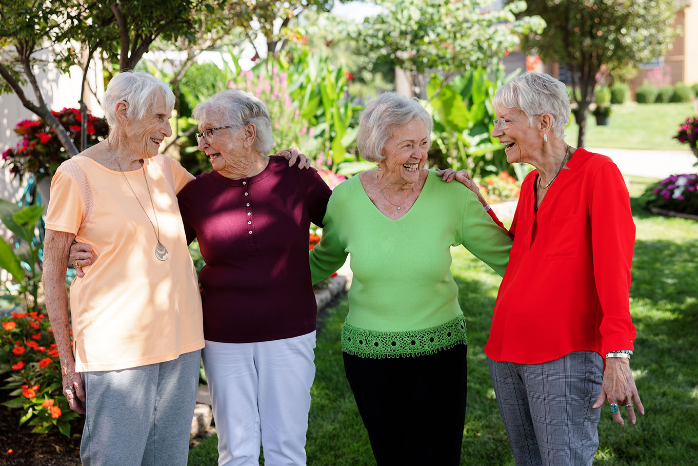 Four smiling residents stand arm in arm in a colorful garden at The Madison Senior Living.