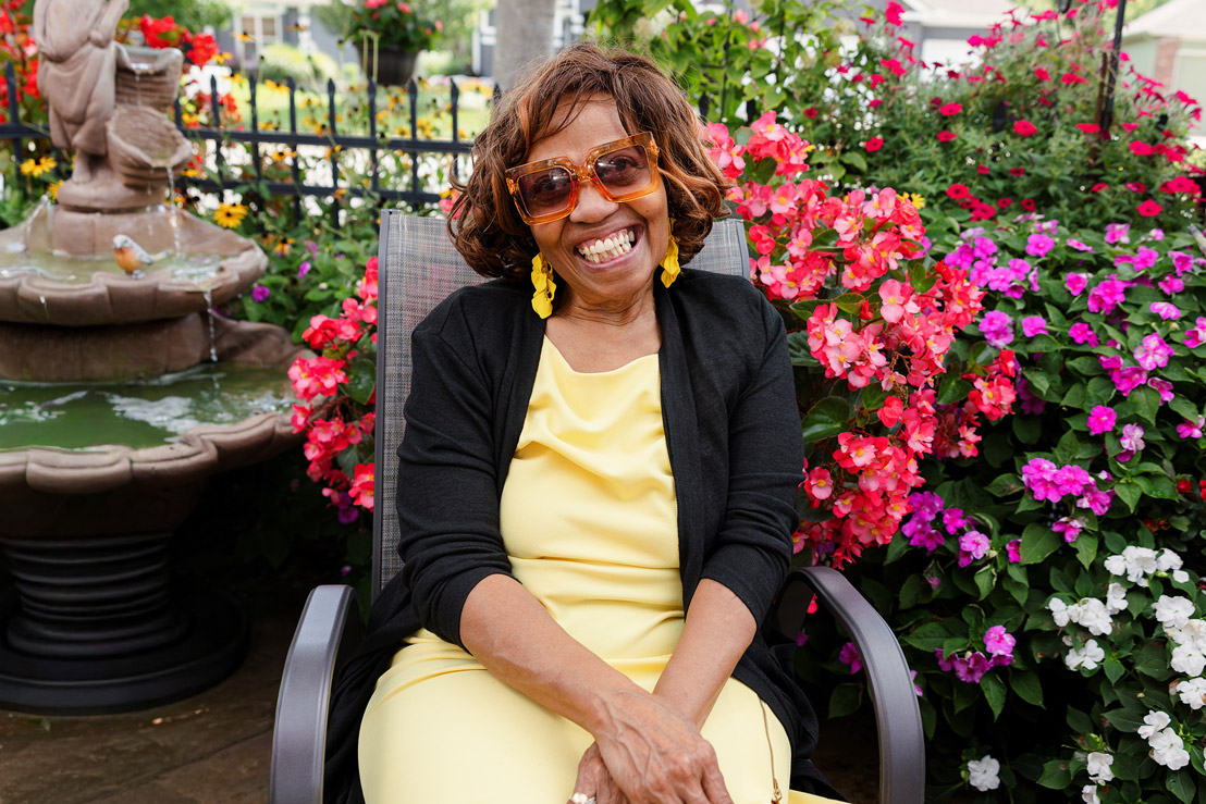 Smiling resident in a yellow dress sits beside a fountain and vibrant flowers at The Madison Senior Living.