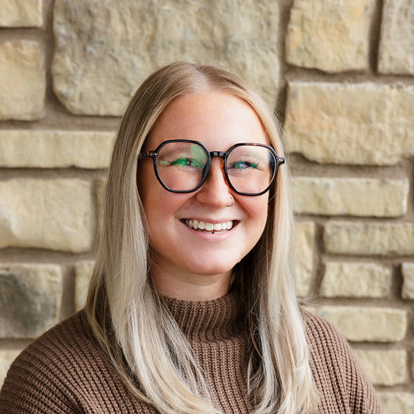 Brittny Middleton, Memory Care Director at The Madison Senior Living, smiling in a professional headshot with long straight blonde hair, oversized glasses, a brown sweater, and a light stone wall background.